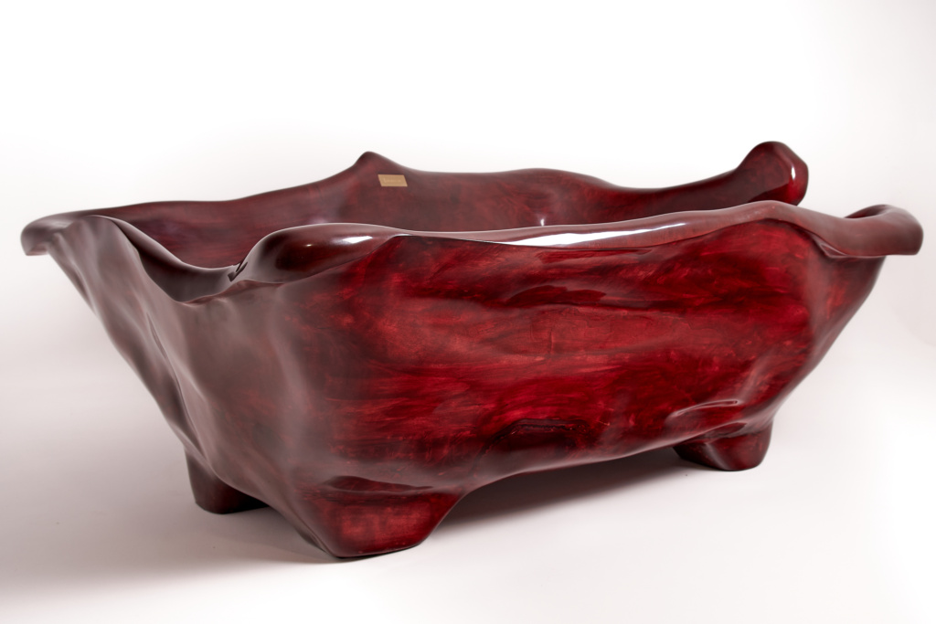 A sculptural bathtub made of polished, deep red wood with smooth, organic curves and irregular edges. The bath has a unique artistic design, suggesting a handmade, custom-crafted piece. 