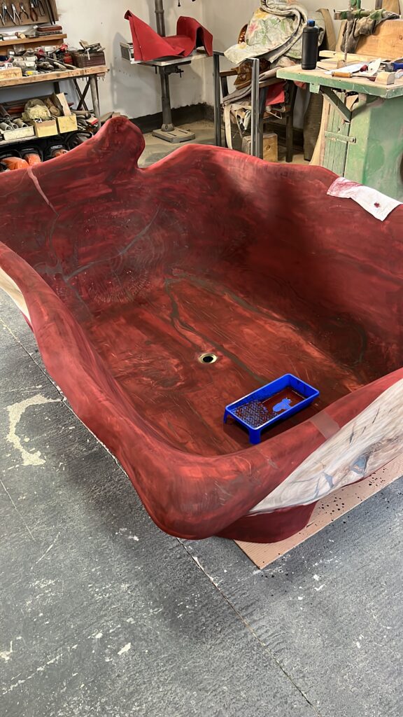 Large, deep, red-toned hand-carved wooden bathtub with organic, uneven edges sits in a workshop; work tools and supplies are visible in the background, and a blue paint tray rests inside the sculptural wood furniture piece.