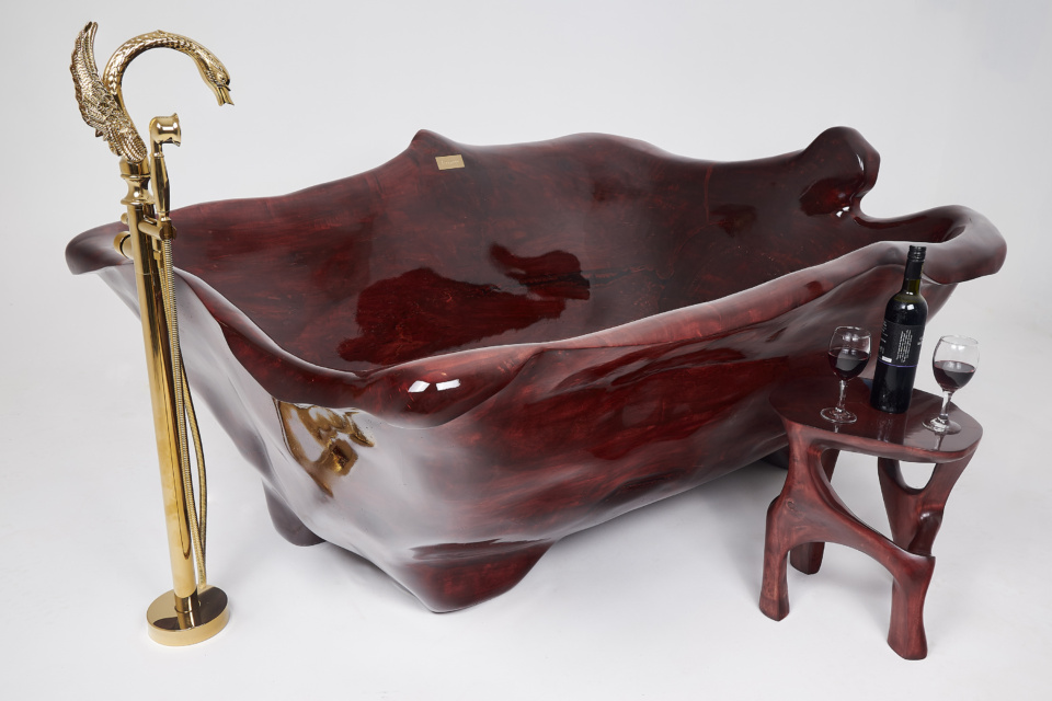 A luxurious, intricately carved red wooden bathtub is paired with a gold swan-shaped faucet. Beside it is a matching wooden table holding a bottle of red wine and two filled glasses, set against a plain white background.