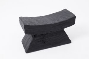 A solid matte black low stool or headrest set against a white background, featuring visible wood grain texture. The piece has a gently curved, concave rectangular top supported by a tapered, trapezoidal block base.